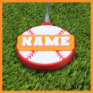 May include: A baseball-themed luggage tag. The tag has the word "NAME" in white block letters on an orange background. The tag is round, with a red border and white center, featuring red stitching details.