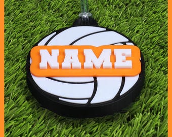 May include: A volleyball-themed ornament with a black, white, and orange design. The ornament features the word "NAME" in white block letters on an orange background, set against a volleyball pattern.