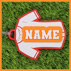 May include: A white and red luggage tag in the shape of a jersey, featuring the word "NAME" in white block letters on an orange rectangle. It has a clear plastic loop for attachment.