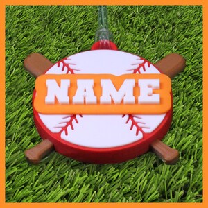 May include: A baseball-themed ornament with a red and white baseball design, featuring the word "NAME" in white letters on an orange background. Two brown bats are positioned on either side of the baseball. The ornament is on a green surface.