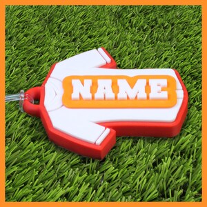 May include: A red, white, and orange luggage tag shaped like a t-shirt. The tag features the word "NAME" in white block letters on an orange rectangle. The tag is on a green surface.