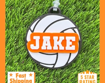 May include: White volleyball ornament with a black outline and an orange stripe across the middle. The name "JAKE" is written in white letters on the orange stripe.