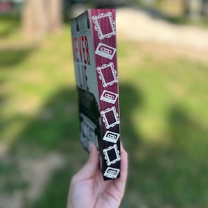 May include: A rectangular wooden sign with the words "NEVER LIE" in red and white lettering. The side of the sign is burgundy with white cassette tape and frame designs. The sign is held in a hand against a blurred green background.