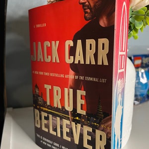 May include: A red, white, and blue paperback book with a gold title, "True Believer" by Jack Carr, the #1 New York Times bestselling author of "The Terminal List". The book cover features a man's face and a cityscape at night.