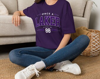 May include: A dark purple short-sleeve t-shirt featuring white and purple text that reads ONCE A LAKER CLASS OF 88. The shirt has a crew neckline and a relaxed fit, paired with blue denim jeans and white sneakers with white laces.