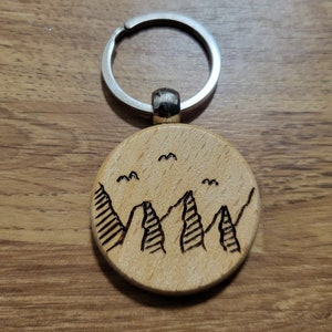 Könnte beinhalten: Ein Holzschlüsselanhänger mit einem runden Design, das eine Bergkette und Vögel im Flug zeigt. Der Schlüsselanhänger ist an einem silbernen Metallring befestigt.