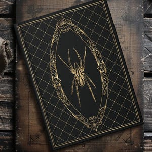 May include: A black journal with a gold spider illustration inside an ornate frame. The cover features a diamond pattern and gold trim. The journal is set on a wooden surface.