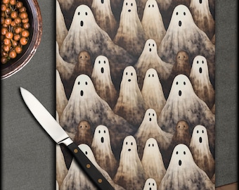 May include: A rectangular cutting board with a repeating pattern of cartoon ghosts in shades of white and brown. A stainless steel knife with a black handle is on the left side of the board. The board is on a gray surface.