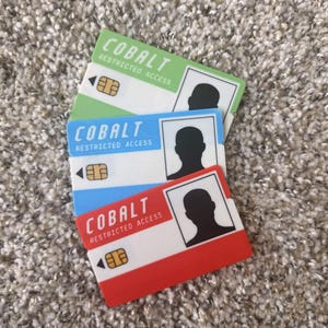 May include: Three rectangular cards with the word "COBALT" and "RESTRICTED ACCESS" printed on them. The cards are green, blue, and red, each with a white border and a silhouette of a person. A gold-coloured chip is on each card.
