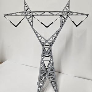 May include: A gray miniature power line tower with a lattice structure. The model features a triangular base and a top section with angled arms, and black insulators. The design is a detailed representation of an electricity pylon.