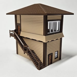 May include: A miniature model of a two-story train station, featuring a brown shingled roof and exterior trim. The building has a light tan siding with a staircase leading to a small balcony. A window with vertical bars is visible.