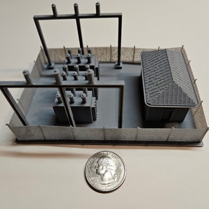 May include: A gray 3D printed model of a small electrical substation with a chain link fence, two transformers, and a small building. A US quarter is shown for scale.
