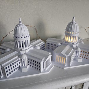 May include: Two white 3D printed models of the Wisconsin State Capitol building. The models are lit from within, creating a warm glow. The models are on a white shelf.
