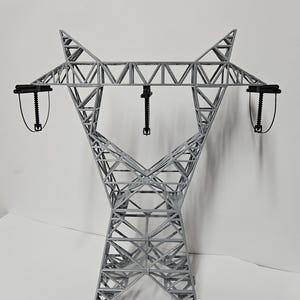 May include: A gray, 3D-printed model of a power line tower. The tower has a lattice structure with two horizontal arms and black insulators. The model is set against a white background.