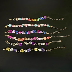 May include: Six beaded flower bracelets in various colors, including pink, blue, orange, and purple. Each bracelet features a gold-colored chain and clasp. The bracelets are arranged on a black surface.