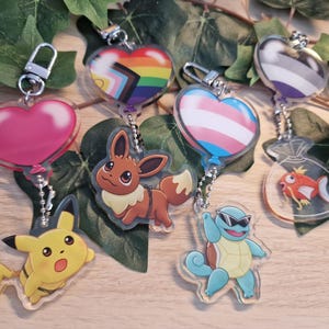 Könnte beinhalten: Vier Acryl-Schlüsselanhänger mit Zeichentrickfiguren und herzförmigen Luftballons. Zu sehen sind Pikachu mit einem rosa Ballon, Evoli mit einem Progress-Pride-Flaggen-Ballon, Schiggy mit einem Transgender-Flaggen-Ballon und Karpador mit einem Asexualitäts-Flaggen-Ballon.