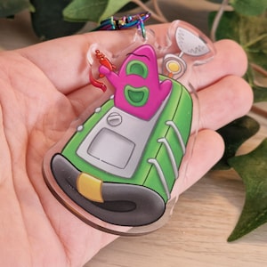 Könnte beinhalten: Ein klarer Acryl-Schlüsselanhänger mit einer Cartoonfigur in einem grün-grauen Gerät. Die Figur ist rosa mit grünen Akzenten und hält eine rote Strahlenpistole. Der Schlüsselanhänger hat einen silberfarbenen Verschluss.