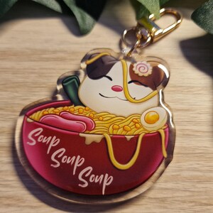 Könnte beinhalten: Transparenter Acryl-Schlüsselanhänger mit einer Cartoon-Katze in einer Ramen-Schüssel. Die rote Schüssel trägt dreimal das Wort „Soup“. Das Ramen enthält Nudeln, Würstchen und ein Ei. Ein goldfarbener Verschluss ist angebracht.