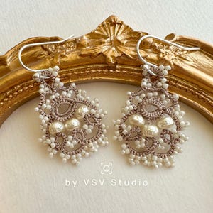 Puede incluir: Un par de aretes delicados con un diseño de crochet marrón y blanco y detalles de perlas. Los aretes cuelgan de ganchos plateados. "by VSV Studio" está escrito debajo de los aretes.