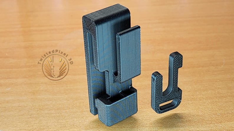 May include: A black plastic 3D printed phone holder with a separate black plastic hook. The holder has a textured surface and a slot for a phone.