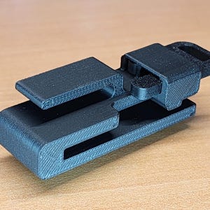 May include: A black plastic clip with a textured surface. The clip has a small loop on the top and a slot on the side.