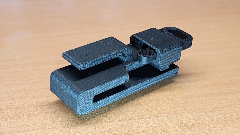 May include: A black plastic clip with a textured surface. The clip has a small loop on the top and a slot on the side.