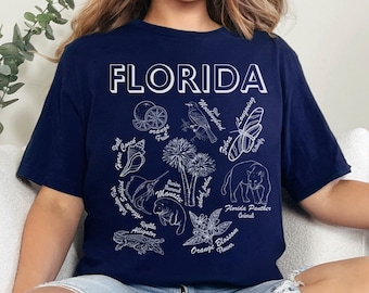May include: A navy blue t-shirt with a white graphic design featuring the word "FLORIDA" and illustrations of various Florida-themed elements, including an orange blossom, a butterfly, a bird, and a seashell.