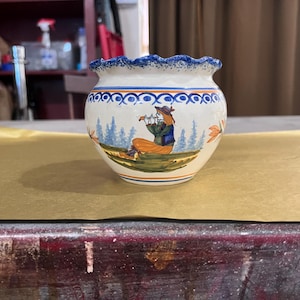Könnte beinhalten: Kleiner, runder Keramiktopf mit einem blau gewellten Rand und einem handgemalten Design. Der Topf zeigt eine sitzende Figur mit Hut, umgeben von Bäumen und Blumen. Der Topf ist weiß mit blauen, orangefarbenen und grünen Akzenten.