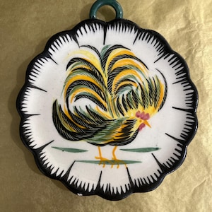 May include: A decorative ceramic plate featuring a colorful rooster design. The plate has a white background with a black scalloped edge. The rooster has yellow, black, and green feathers. A green loop is at the top for hanging.