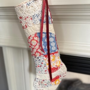 May include: A patchwork Christmas stocking with a red velvet ribbon and white crocheted trim. The stocking is made of various fabrics in shades of red, blue, and white with floral patterns.