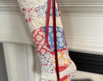 May include: A patchwork Christmas stocking with a red velvet ribbon and white crocheted trim. The stocking is made of various fabrics in shades of red, blue, and white with floral patterns.