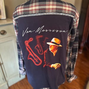 May include: A navy blue flannel shirt with a plaid pattern on the shoulders and sleeves. The back features a graphic of Van Morrison with a saxophone and the artist's signature in white script. A unique, music-themed apparel item.