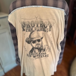 Puede incluir: Camiseta de manga larga color beige con un gráfico de un vaquero con gafas de sol y el texto "PROTECT THE BRAND" y "RANCH HAND RIP WHEELER Wrangler". La camisa tiene un estampado de cuadros en los hombros y la parte superior de la espalda.