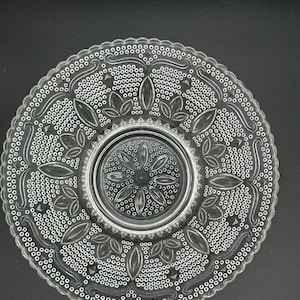Puede incluir: Un plato de vidrio transparente con un patrón decorativo intrincado de pequeños círculos y espirales. El plato tiene una sección central elevada.