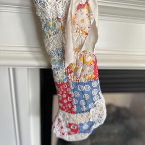 May include: A patchwork Christmas stocking with a white crocheted trim and a white ribbon bow. The stocking is made of various floral and geometric fabrics in red, blue, and yellow.