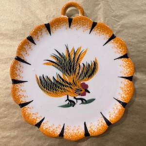 May include: A ceramic plate with a scalloped edge and a loop for hanging. The plate features a rooster illustration in shades of yellow, orange, black, and green on a white background. The rim is orange with black accents.