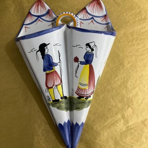 May include: White ceramic wall pocket with a hand-painted design. The pocket features a split design with a handle and a painted scene of two figures in traditional clothing. The top has red and blue accents, and the bottom is blue.