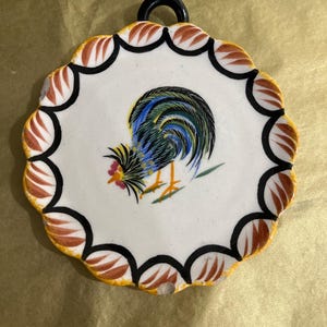 May include: A decorative ceramic rooster trivet. The trivet features a white center with a colorful rooster illustration. The scalloped edge is white, black, and brown. A black loop is at the top for hanging.
