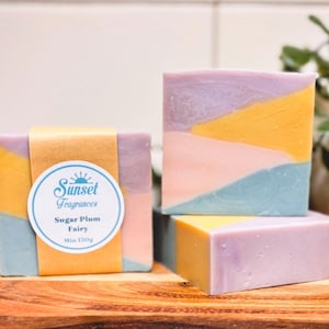 May include: Handmade soap bars in pastel shades of yellow, blue, pink, and purple. One bar is wrapped with a paper band and a label that reads "Sunset Fragrances Sugar Plum Fairy Min 134g."
