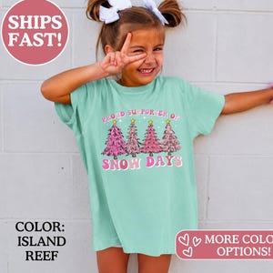 May include: A light green short-sleeve t-shirt featuring a graphic of four pink Christmas trees with yellow stars. The text reads PROUD SUPPORTER OF SNOW DAYS in pink and white lettering. The shirt has a crew neckline and a relaxed fit.