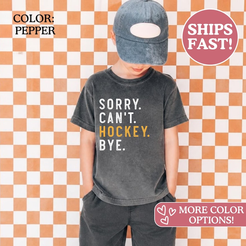 May include: A pepper-colored short-sleeve t-shirt featuring the white and yellow text SORRY. CAN'T. HOCKEY. BYE. The shirt is paired with a matching pepper-colored baseball cap with a blank front panel, creating a coordinated casual outfit.