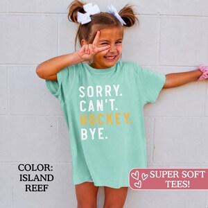 May include: A light green short-sleeve t-shirt featuring the white text SORRY. CAN'T. and the yellow text HOCKEY. followed by the white text BYE. The shirt has a crew neckline and a relaxed fit, with the color identified as Island Reef.