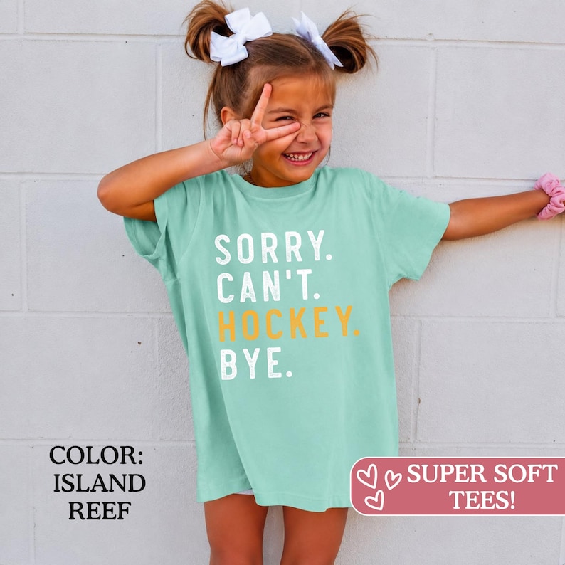 May include: A light green short-sleeve t-shirt featuring the white text SORRY. CAN'T. and the yellow text HOCKEY. followed by the white text BYE. The shirt has a crew neckline and a relaxed fit, with the color identified as Island Reef.