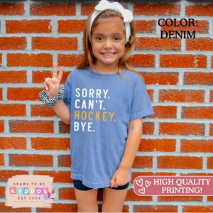 May include: A denim blue short-sleeve t-shirt featuring the white and yellow text SORRY. CAN'T. HOCKEY. BYE. printed on the front. The shirt has a crew neckline and a relaxed fit, showcasing a casual style with bold, clear lettering across the chest.