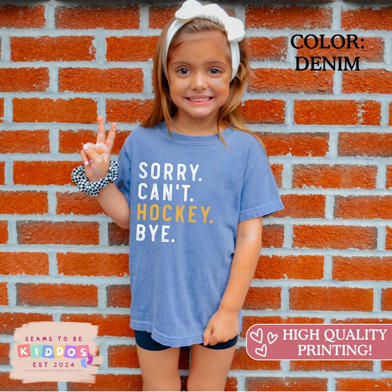 May include: A denim blue short-sleeve t-shirt featuring the white and yellow text SORRY. CAN'T. HOCKEY. BYE. printed on the front. The shirt has a crew neckline and a relaxed fit, showcasing a casual style with bold, clear lettering across the chest.