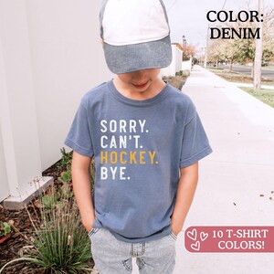 May include: A denim blue short-sleeve t-shirt featuring the white and yellow text SORRY. CAN'T. HOCKEY. BYE. The shirt is paired with a two-tone baseball cap with a white front panel and a denim blue brim. The shirt has a crew neckline.