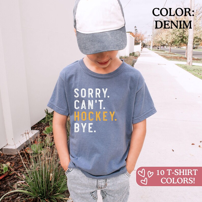 May include: A denim blue short-sleeve t-shirt featuring the white and yellow text SORRY. CAN'T. HOCKEY. BYE. The shirt is paired with a two-tone baseball cap with a white front panel and a denim blue brim. The shirt has a crew neckline.