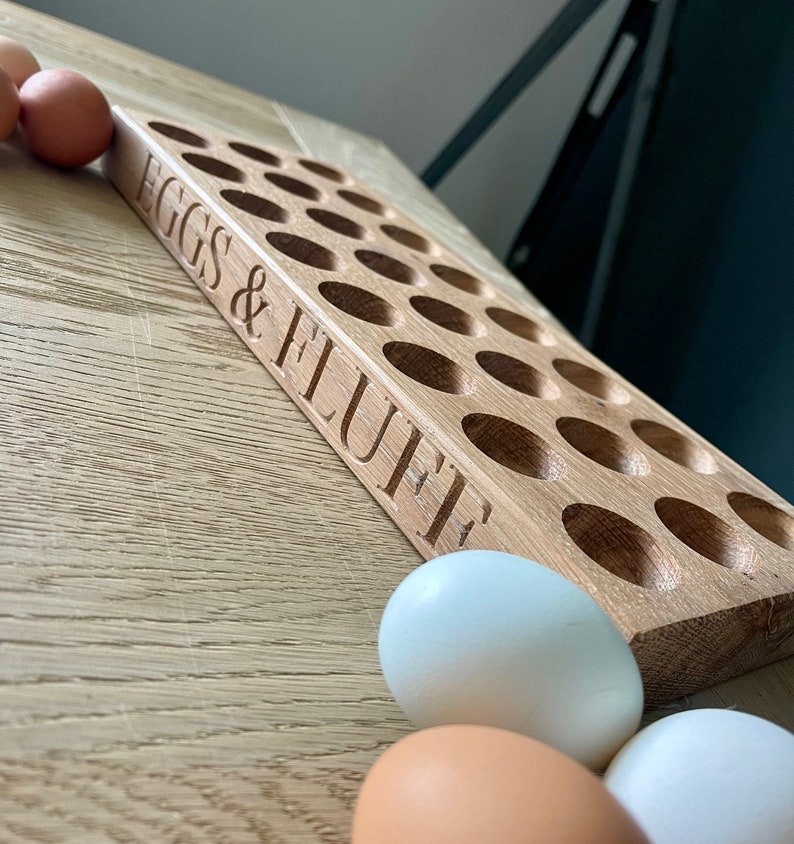 May include: A wooden egg holder with 24 slots, labeled "EGGS & FLUFFE". Three white eggs are in the foreground.