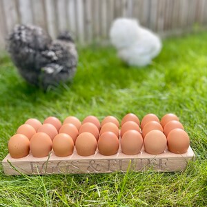 May include: A wooden egg crate with the words "Eggs & Fluff" engraved on the front. The crate is filled with brown eggs and is sitting on a bed of green grass.