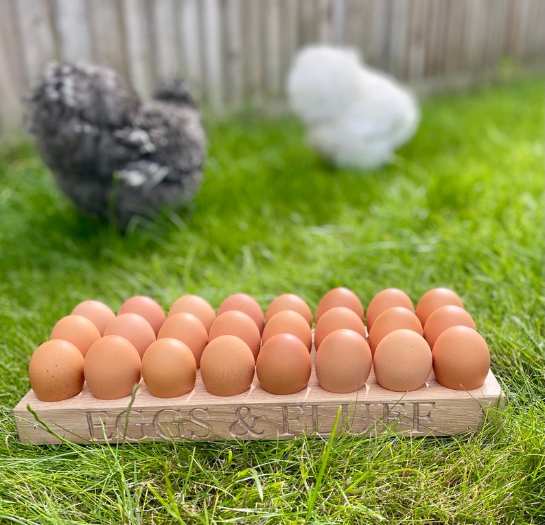 May include: A wooden egg crate with the words "Eggs & Fluff" engraved on the front. The crate is filled with brown eggs and is sitting on a bed of green grass.